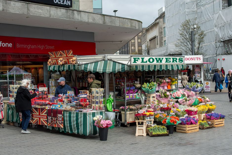 A flower stall near Wood Green station displays an array of fresh, vibrant floral arrangements and bouquets wrapped in colorful paper and cellophane, featuring a variety of flowers including roses, tu