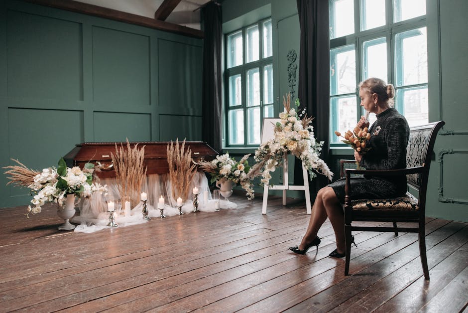 A woman with gray hair styled in a bun, dressed in a black lace dress and black heels, is seated on a vintage wooden chair with a cushioned seat in a softly lit room with large green painted walls and