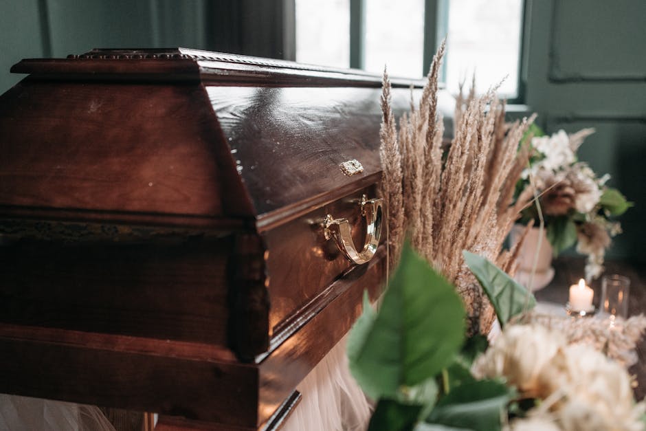 A polished dark brown wooden casket adorned with gold handles, situated in a softly lit indoor setting. Surrounding the casket are elegant floral arrangements featuring white and blush peonies, and va