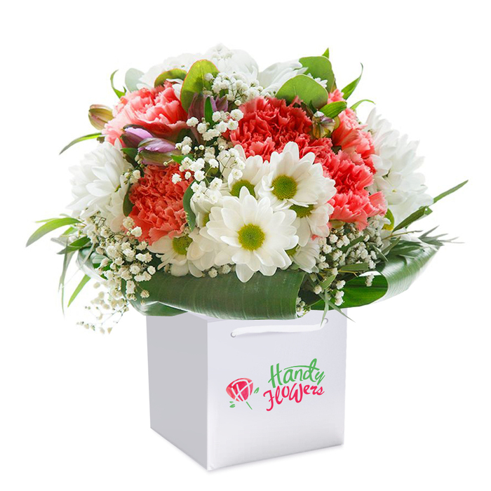 Carefully arranged within a modern square white gift bag, this radiant bouquet offers a luminous statement of joy and affection, artfully composed in the heart of Wood Green. Cascading coral-pink and peach carnations-rich with ruffled, layered petals-draw the eye inward, their warmth amplified by the gentle gleam of natural light often found in the open spaces near Woodside Park. Framing their vigour are crisp white daisy-like chrysanthemums with vivid yellow-green centres, each bloom adding a spirit of cheer akin to Wood Green's welcoming high street. Delicate sprigs of snowy white baby's breath are scattered throughout, their gossamer blossoms providing breath and softness between the vibrant main flowers like a cool morning breeze. A collar of glossy, deep green leaves fans outward, embracing the arrangement and imparting a sense of occasion-both protective and decorative, echoing the tranquil green spaces that punctuate the neighbourhood. Subtle, unopened buds with hints of purple and pink are tucked into the bouquet, introducing intrigue and depth, enhancing the arrangement's naturalistic charm. The white bag, designed for easy gifting, sits ready for delivery to any address in Wood Green or nearby Harringay, clearly highlighting the freshness and expert craftsmanship behind each order. The entire image is set against a spotless white background, harmonizing with the clean, contemporary spirit of local floristry and offering a visual celebration perfect for marking birthdays, anniversaries, or sending an uplifting gesture to a loved one close by.